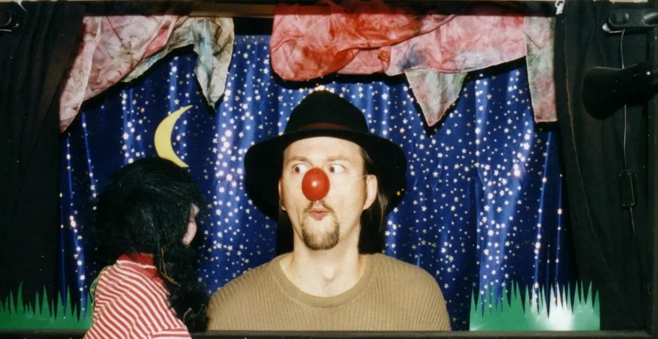 Ein Mann mit Hut und roter Clownsnase schaut aus einem Puppentheater. Eine Handpuppe steht davor und schaut ihn an.