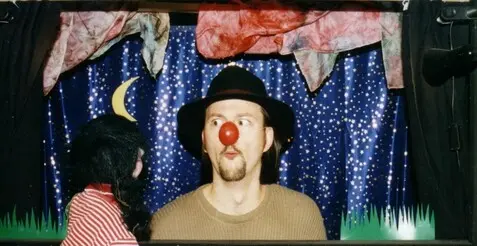 Ein Mann mit Hut und roter Clownsnase schaut aus einem Puppentheater. Eine Handpuppe steht davor und schaut ihn an.